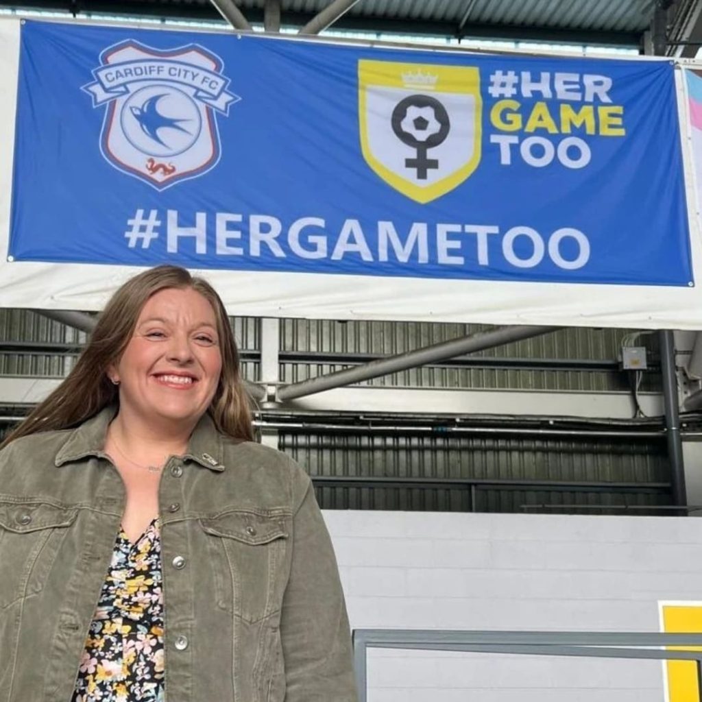 Photograph of Cardiff City FC's Her Game Too ambassador Brittany Laing. She is smiling below a Cardiff City Bluebirds Her Game Too flag