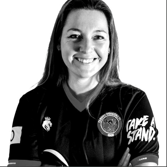 Grayscale Photograph of Founder Charlotte Galloway, smiling, wearing Cardiff Dragons FC black women's football shirt, made by Hope & Glory Sportswear. Take a Stand logo on left sleeve