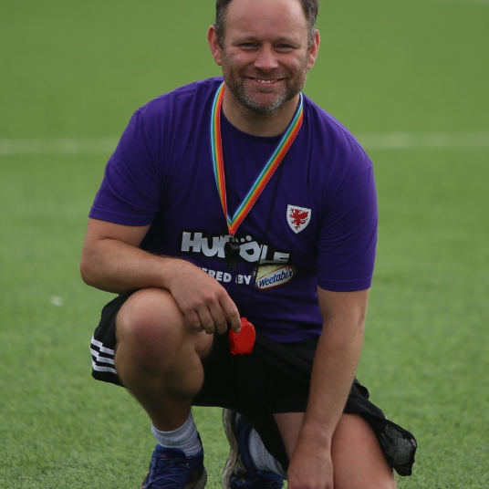 Photograph of Founder and Coach Ryan McGavock. Kneeling on football pitch at the Cardiff City Ladies FC Huddle Unite festival at Ocean Park Arena. Wearing a whistle around his neck with a rainbow pride lanyard. Smiling