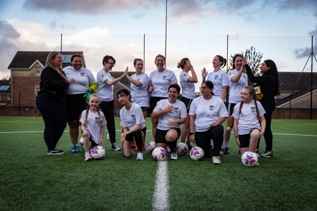 Group photo of womens football training session. 10 women and 2 girls wearing white "Her Game Too" t-shirts
